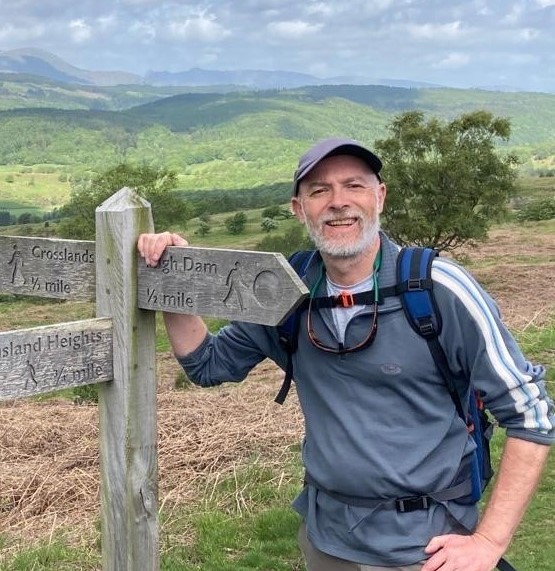 Jerry Cripps, Chartered Counselling Psychologist, standing at a countryside signpost with hills in the background